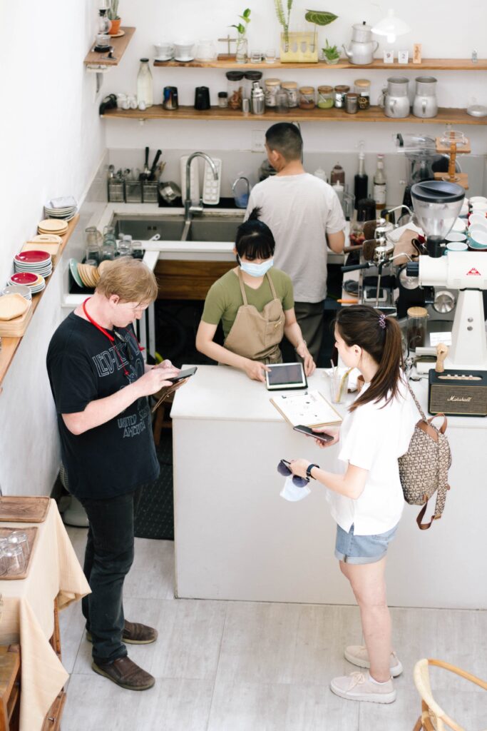 2 beverage service workers behind a counter, 1 of them holding a tablet waiting for one of two customers to pay via their cell phone.